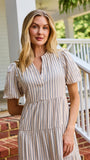 A woman stands on red brick steps in front of a white house porch, wearing a cream and tan vertically striped midi dress with puff sleeves and a tiered ruffle hem.