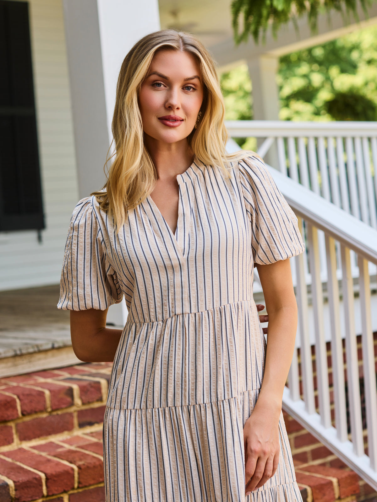 A woman stands on red brick steps in front of a white house porch, wearing a cream and tan vertically striped midi dress with puff sleeves and a tiered ruffle hem.