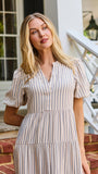 A woman stands on red brick steps in front of a white house porch, wearing a cream and tan vertically striped midi dress with puff sleeves and a tiered ruffle hem.