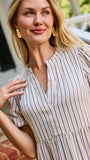 A woman stands on red brick steps in front of a white house porch, wearing a cream and tan vertically striped midi dress with puff sleeves and a tiered ruffle hem.