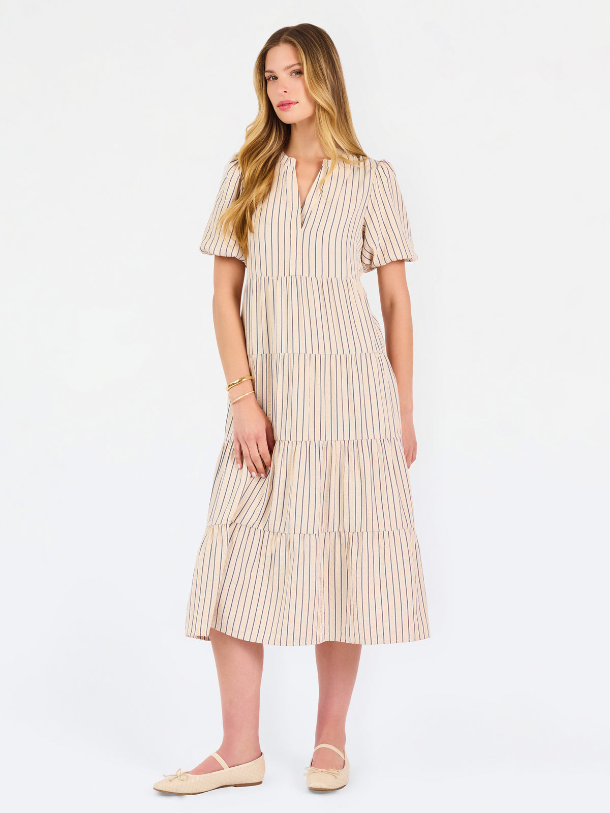 A woman modeling a short-sleeve midi dress with vertical beige and white stripes, featuring a tiered ruffle hem and a split neckline, set against a plain white studio background.