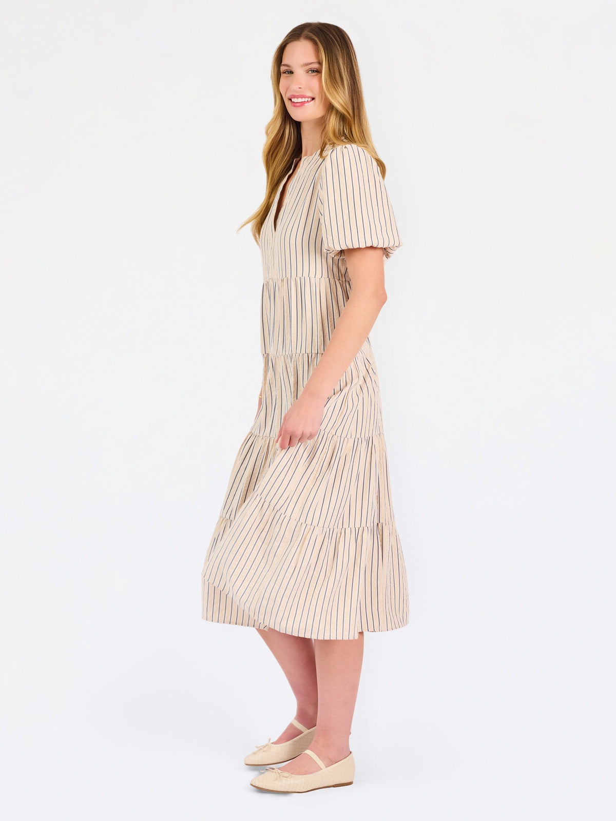 A woman modeling a short-sleeve midi dress with vertical beige and white stripes, featuring a tiered ruffle hem and a split neckline, set against a plain white studio background.