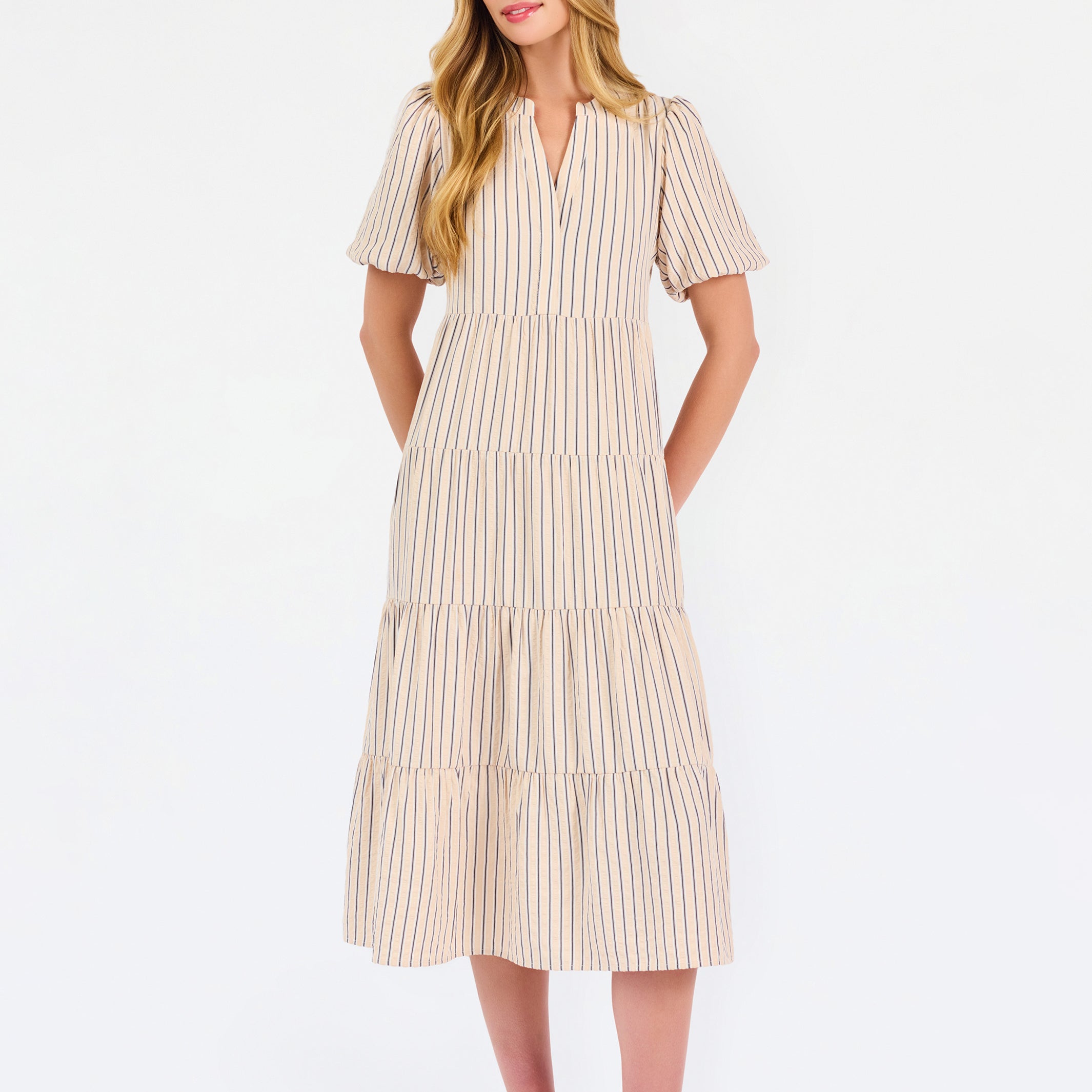 A woman modeling a short-sleeve midi dress with vertical beige and white stripes, featuring a tiered ruffle hem and a split neckline, set against a plain white studio background.