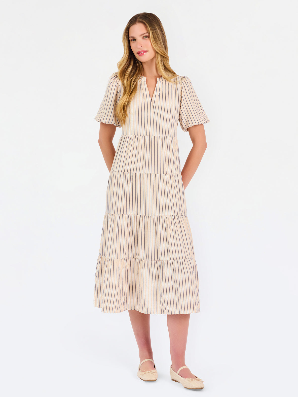 A woman modeling a short-sleeve midi dress with vertical beige and white stripes, featuring a tiered ruffle hem and a split neckline, set against a plain white studio background.