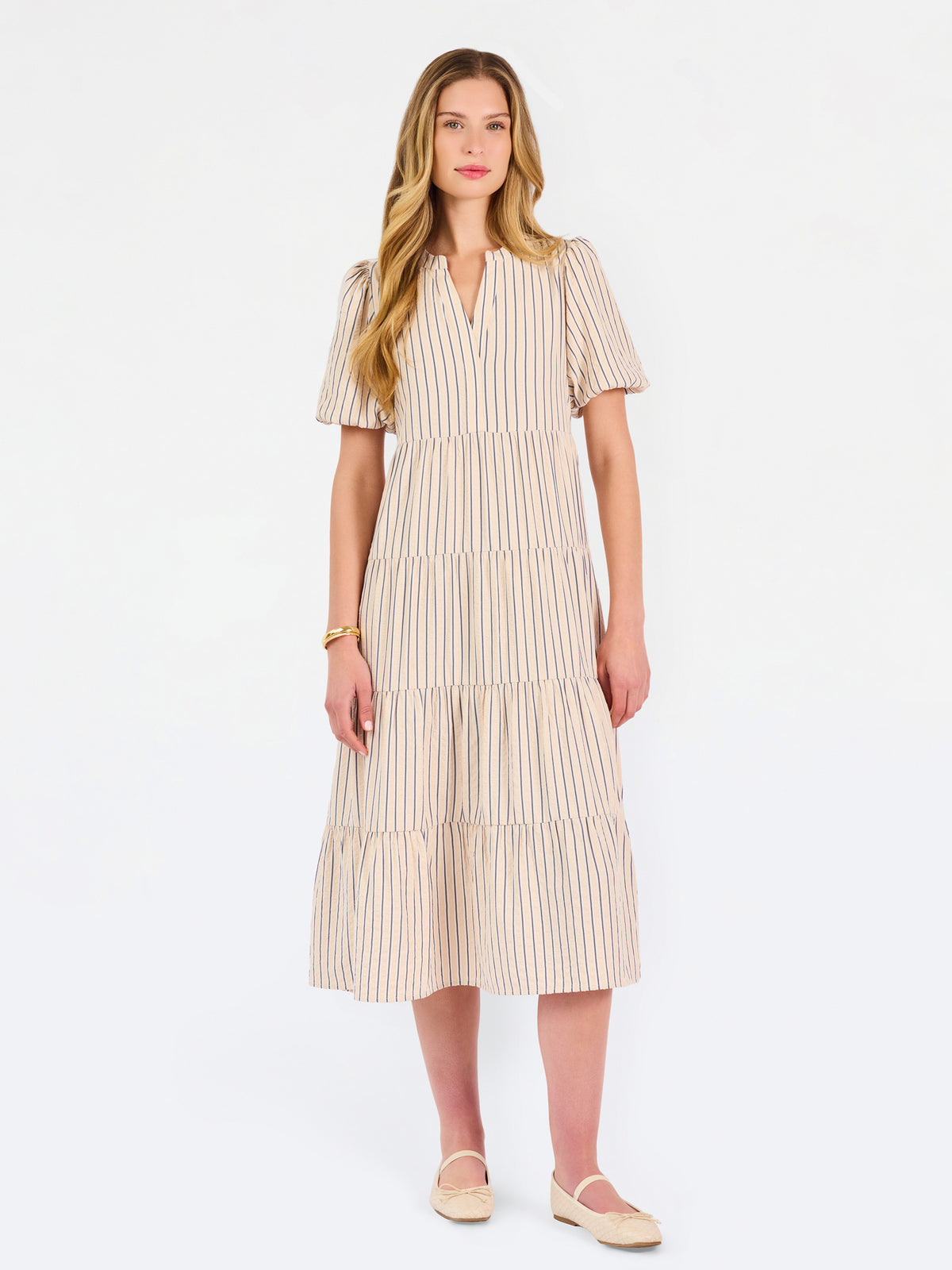 A woman modeling a short-sleeve midi dress with vertical beige and white stripes, featuring a tiered ruffle hem and a split neckline, set against a plain white studio background.