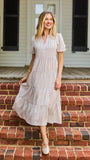 A woman stands on red brick steps in front of a white house porch, wearing a cream and tan vertically striped midi dress with puff sleeves and a tiered ruffle hem.