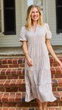 A woman stands on red brick steps in front of a white house porch, wearing a cream and tan vertically striped midi dress with puff sleeves and a tiered ruffle hem.