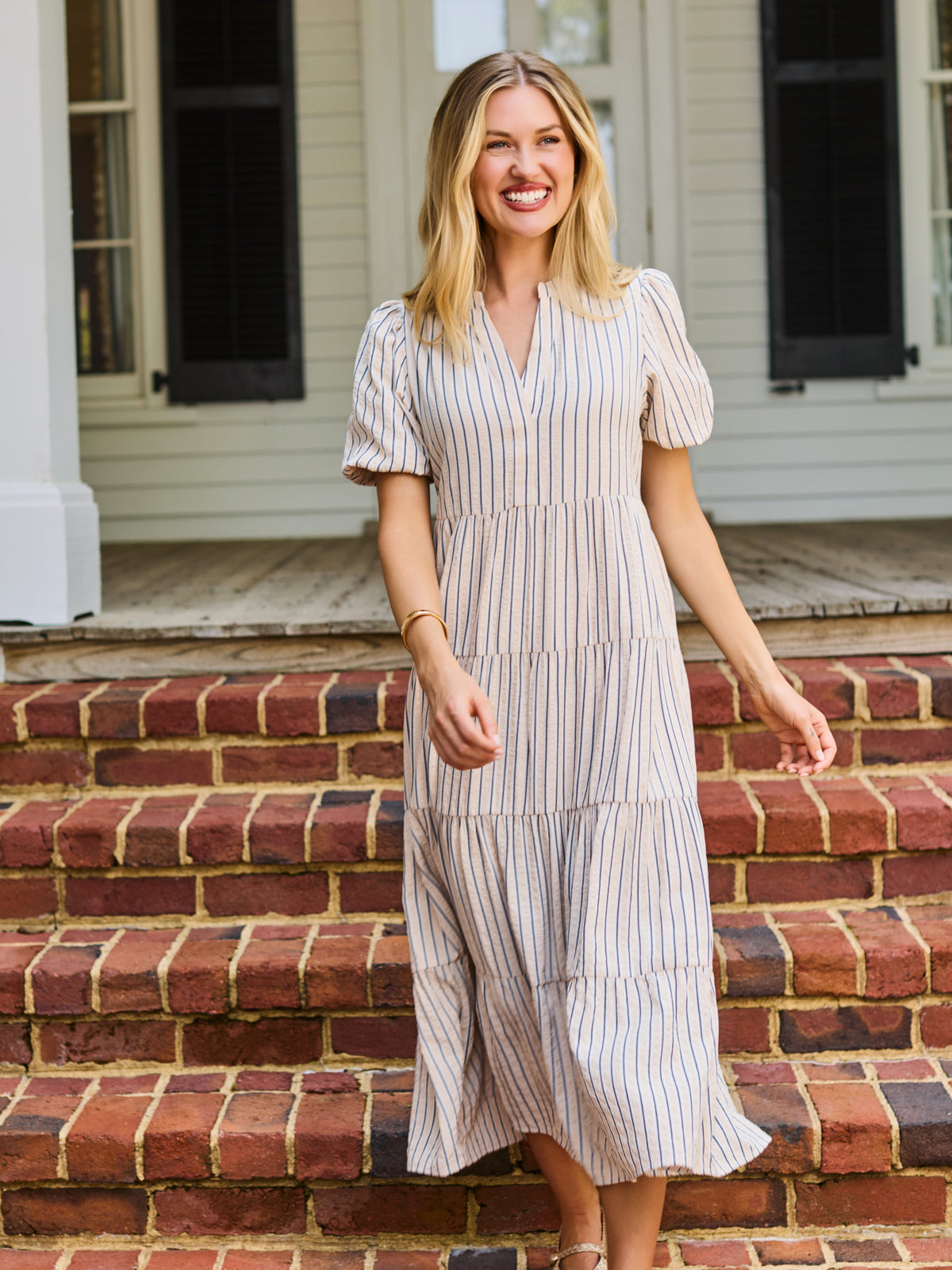 A woman stands on red brick steps in front of a white house porch, wearing a cream and tan vertically striped midi dress with puff sleeves and a tiered ruffle hem.