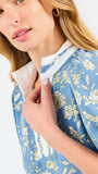 A woman modeling a light blue short-sleeve blouse with a white and yellow floral pattern, featuring a split neckline with white trim and button details, paired with blue jeans against a plain white background.