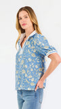 A woman modeling a light blue short-sleeve blouse with a white and yellow floral pattern, featuring a split neckline with white trim and button details, paired with blue jeans against a plain white background.