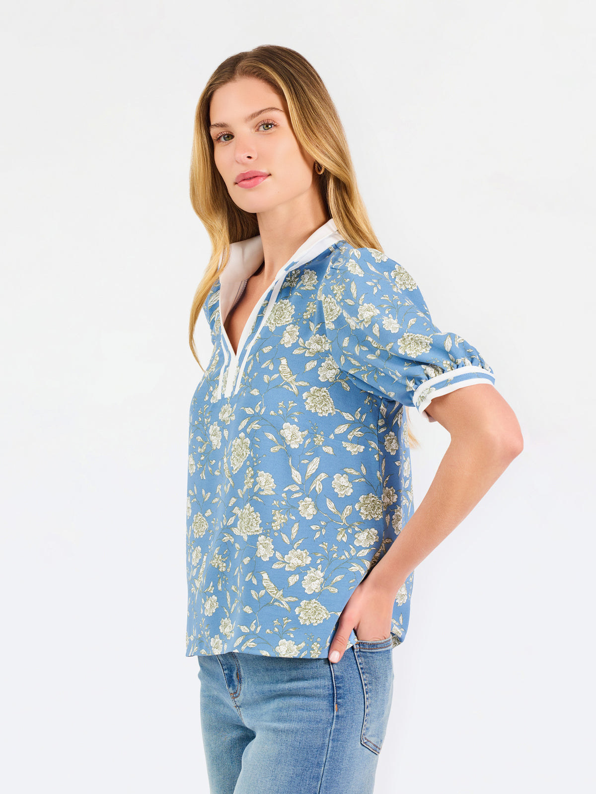 A woman modeling a light blue short-sleeve blouse with a white and yellow floral pattern, featuring a split neckline with white trim and button details, paired with blue jeans against a plain white background.