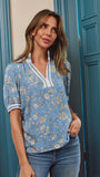 A woman wearing a light blue short-sleeve blouse with a white and beige floral pattern, paired with blue jeans, standing in front of teal paneled doors.