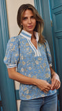 A woman wearing a light blue short-sleeve blouse with a white and beige floral pattern, paired with blue jeans, standing in front of teal paneled doors.