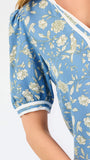 A woman modeling a light blue short-sleeve blouse with a white and yellow floral pattern, featuring a split neckline with white trim and button details, paired with blue jeans against a plain white background.