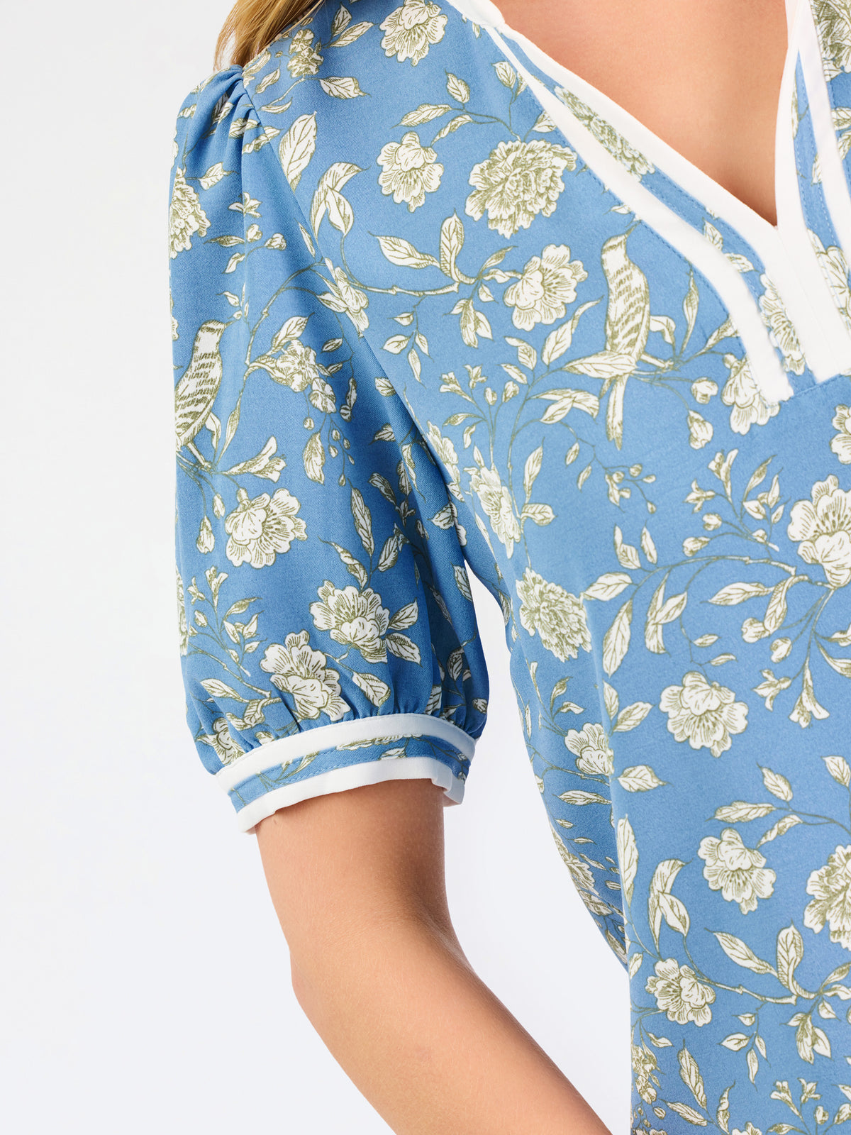 A woman modeling a light blue short-sleeve blouse with a white and yellow floral pattern, featuring a split neckline with white trim and button details, paired with blue jeans against a plain white background.