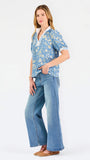 A woman modeling a light blue short-sleeve blouse with a white and yellow floral pattern, featuring a split neckline with white trim and button details, paired with blue jeans against a plain white background.