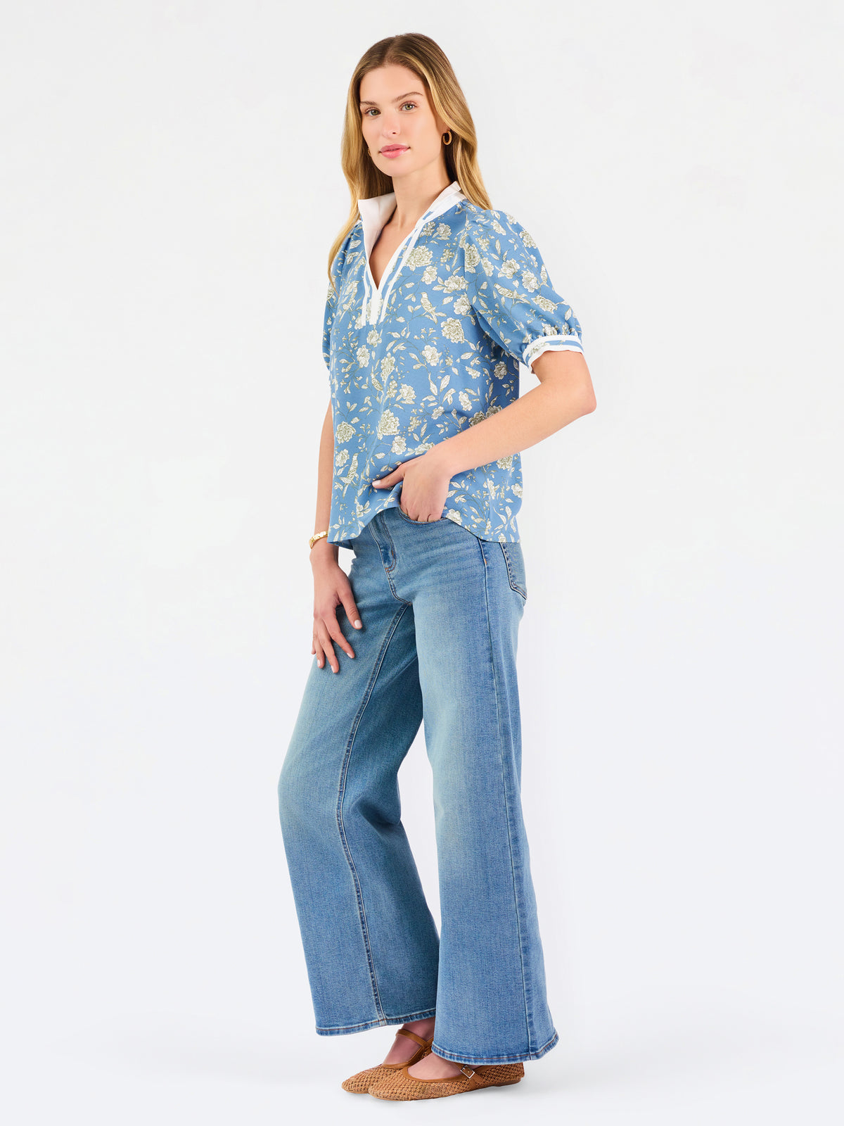 A woman modeling a light blue short-sleeve blouse with a white and yellow floral pattern, featuring a split neckline with white trim and button details, paired with blue jeans against a plain white background.