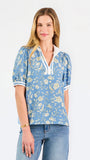 A woman modeling a light blue short-sleeve blouse with a white and yellow floral pattern, featuring a split neckline with white trim and button details, paired with blue jeans against a plain white background.
