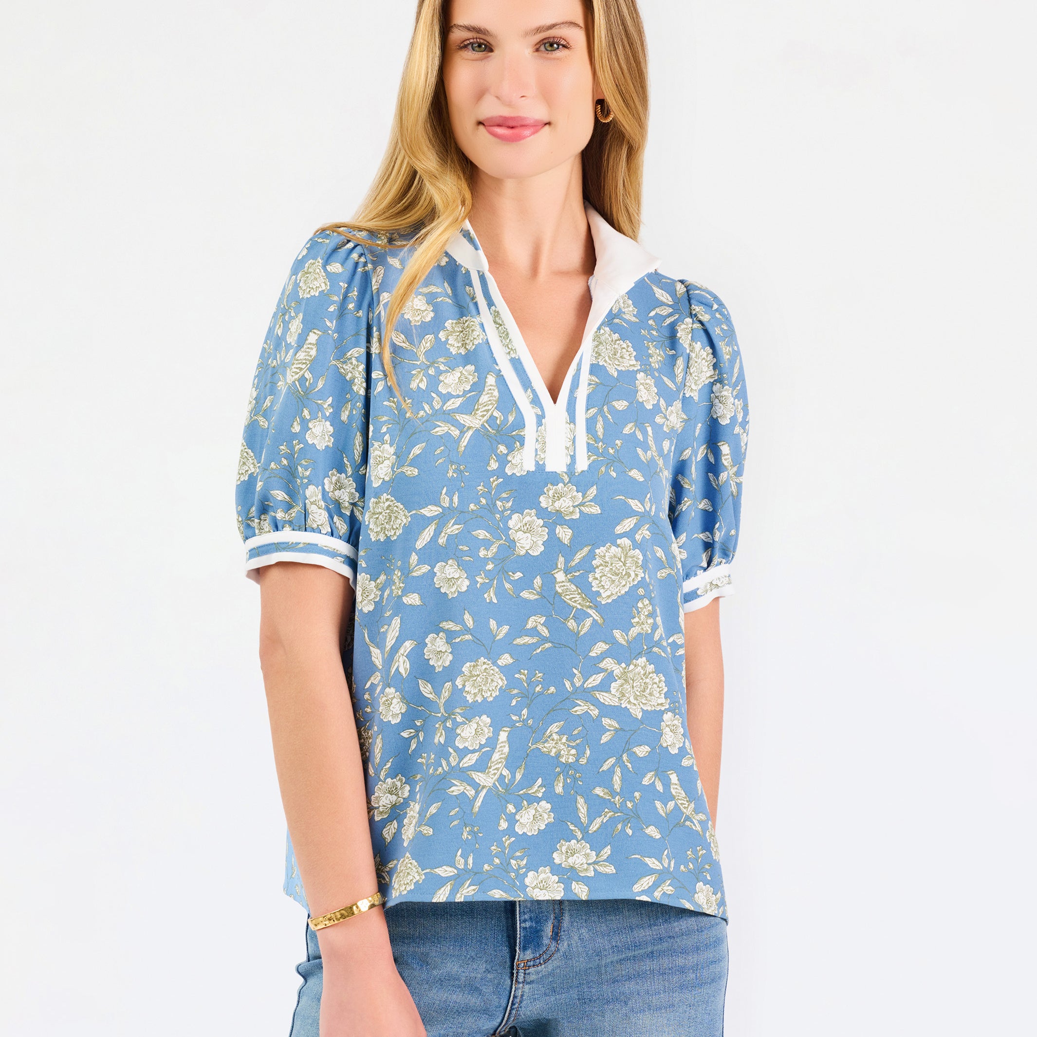 A woman modeling a light blue short-sleeve blouse with a white and yellow floral pattern, featuring a split neckline with white trim and button details, paired with blue jeans against a plain white background.