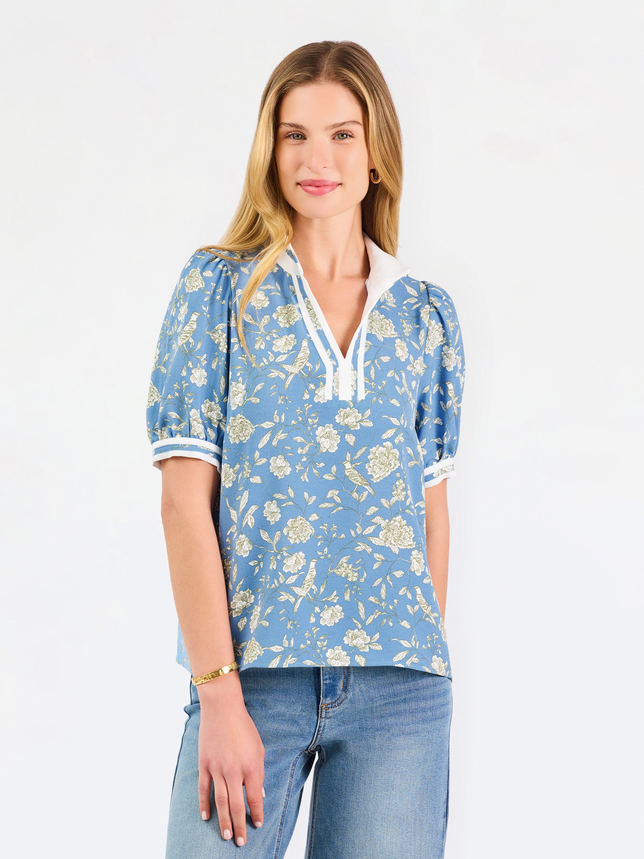 A woman modeling a light blue short-sleeve blouse with a white and yellow floral pattern, featuring a split neckline with white trim and button details, paired with blue jeans against a plain white background.