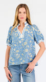 A woman modeling a light blue short-sleeve blouse with a white and yellow floral pattern, featuring a split neckline with white trim and button details, paired with blue jeans against a plain white background.