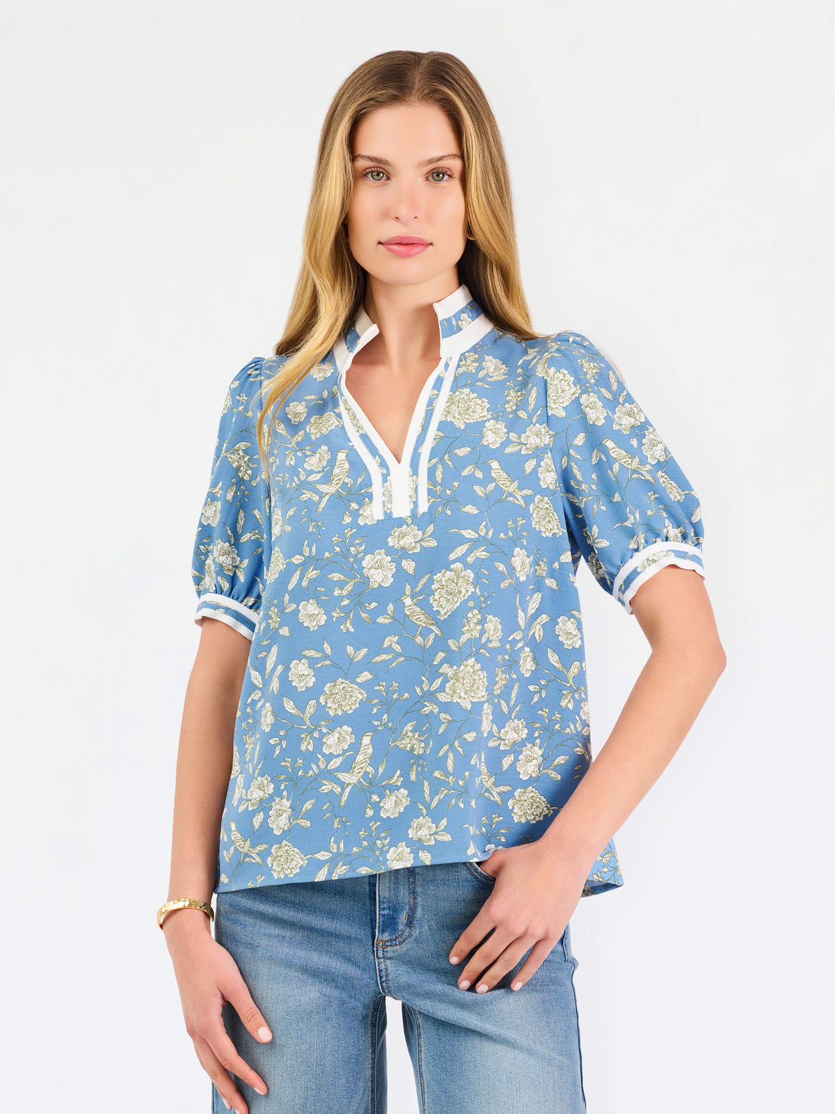 A woman modeling a light blue short-sleeve blouse with a white and yellow floral pattern, featuring a split neckline with white trim and button details, paired with blue jeans against a plain white background.