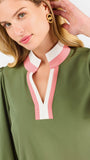 A close-up of a woman wearing an olive green long-sleeve blouse with a split V-neckline featuring pink and white striped trim. She is wearing gold double-ring earrings and touching her hair against a plain light background.