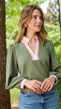 A woman wearing an olive green long-sleeve blouse with a pink and white V-neck placket, paired with blue jeans and a brown belt, standing outdoors next to a tree.