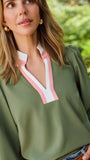 A woman wearing an olive green long-sleeve blouse with a pink and white V-neck placket, paired with blue jeans and a brown belt, standing outdoors next to a tree.