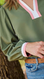A woman wearing an olive green long-sleeve blouse with a pink and white V-neck placket, paired with blue jeans and a brown belt, standing outdoors next to a tree.