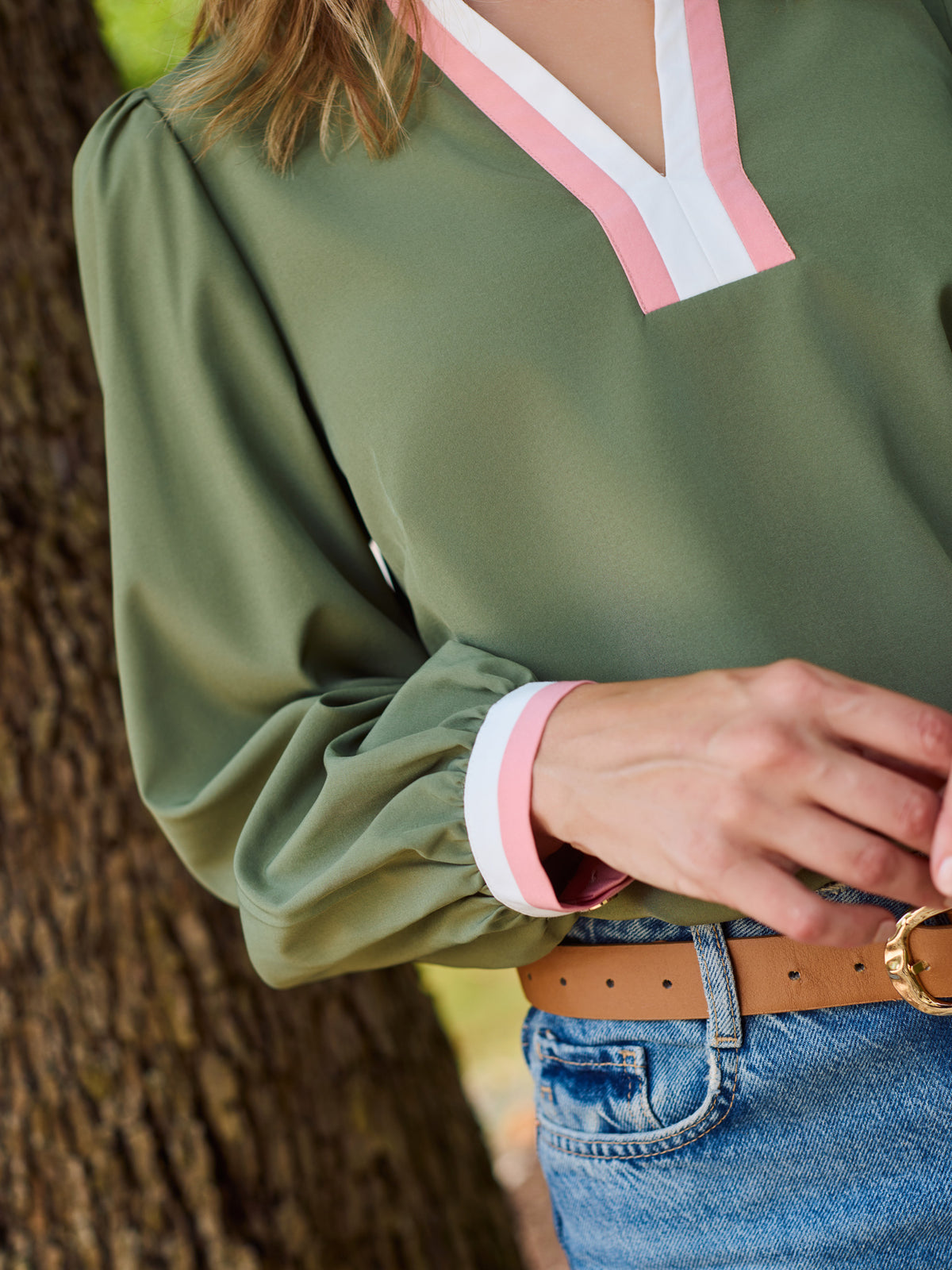 A woman wearing an olive green long-sleeve blouse with a pink and white V-neck placket, paired with blue jeans and a brown belt, standing outdoors next to a tree.