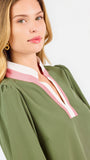 A woman modeling a long-sleeve olive green blouse with a pink and white V-neck trim, paired with blue jeans, against a plain white studio background.