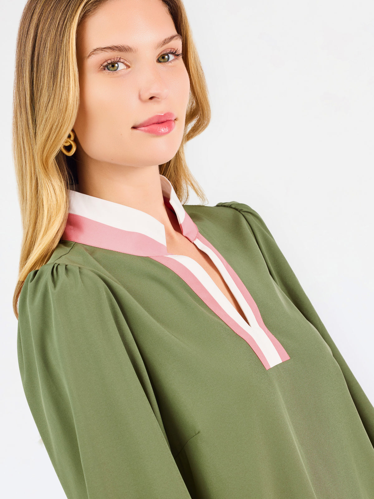 A woman modeling a long-sleeve olive green blouse with a pink and white V-neck trim, paired with blue jeans, against a plain white studio background.