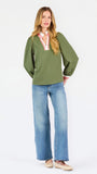 A woman modeling a green long-sleeve blouse with a pink and white striped V-neckline, paired with wide-leg blue jeans and beige shoes, against a plain white studio background.