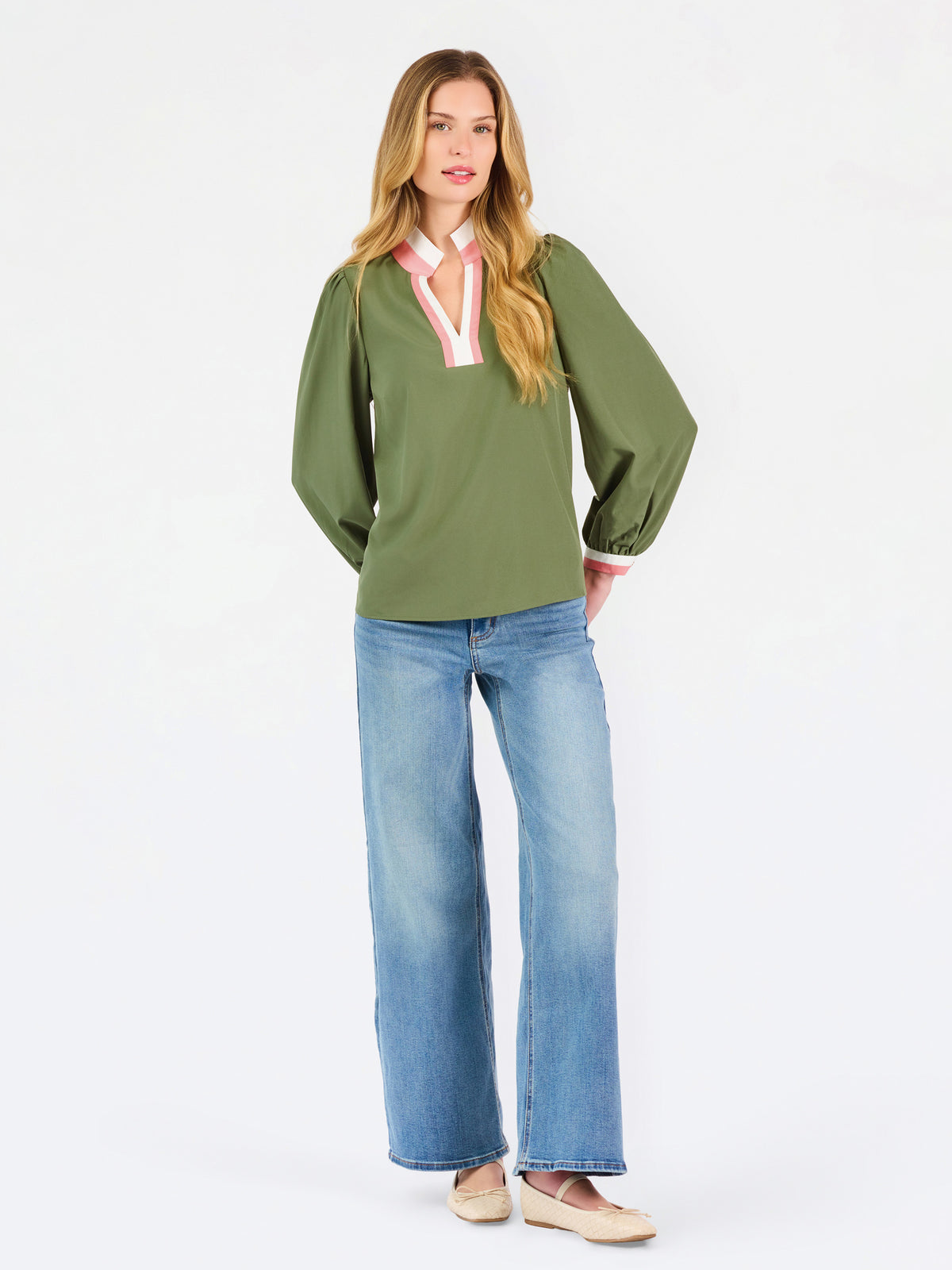 A woman modeling a green long-sleeve blouse with a pink and white striped V-neckline, paired with wide-leg blue jeans and beige shoes, against a plain white studio background.