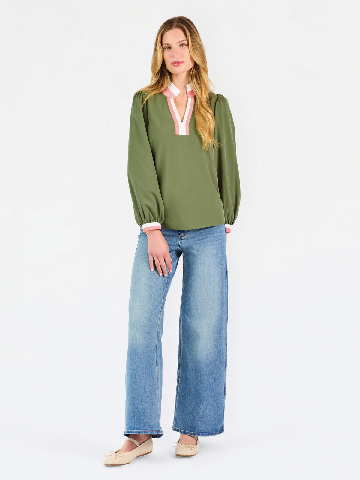 A woman modeling a long-sleeve olive green blouse with a pink and white V-neck trim, paired with blue jeans, against a plain white studio background.