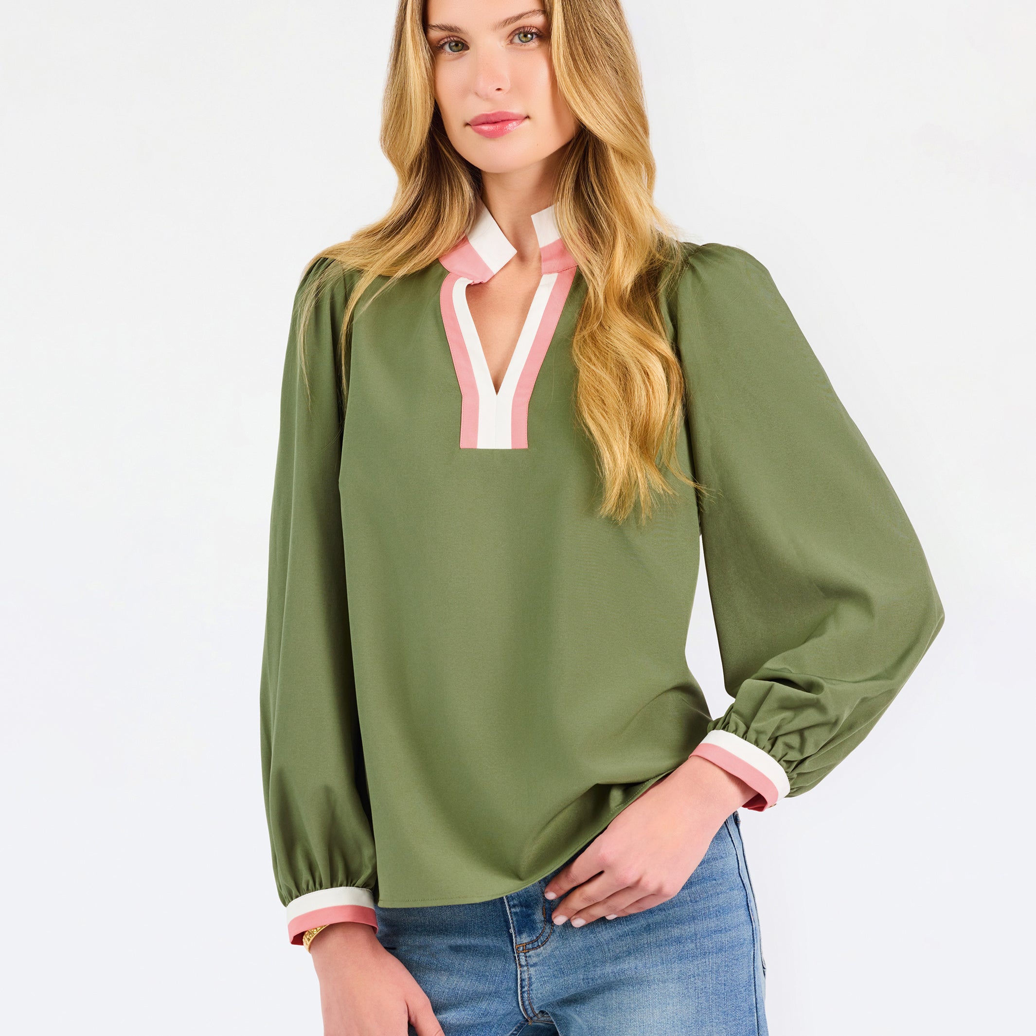 A woman modeling a long-sleeve olive green blouse with a pink and white V-neck trim, paired with blue jeans, against a plain white studio background.