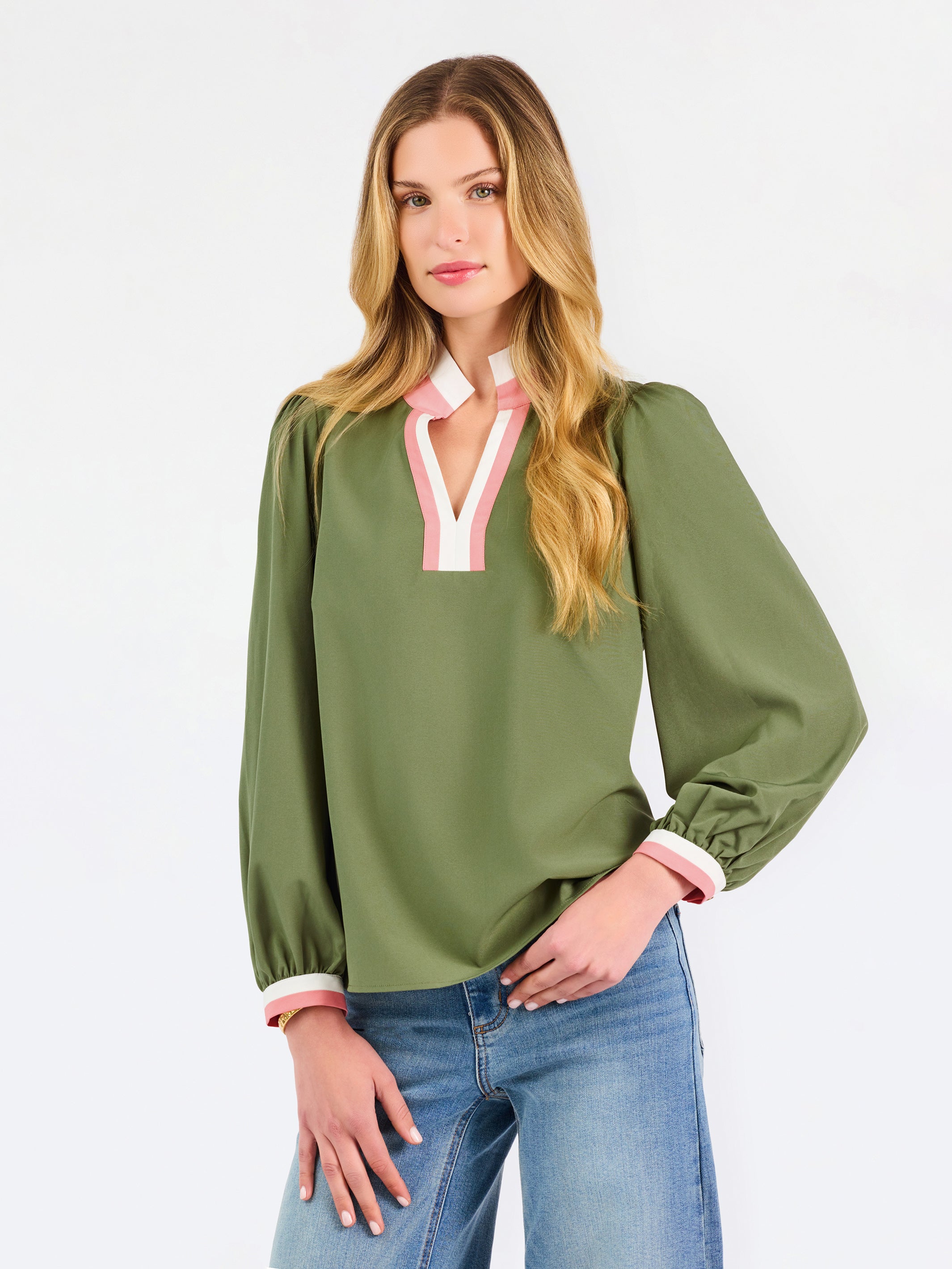 A woman modeling a long-sleeve olive green blouse with a pink and white V-neck trim, paired with blue jeans, against a plain white studio background.