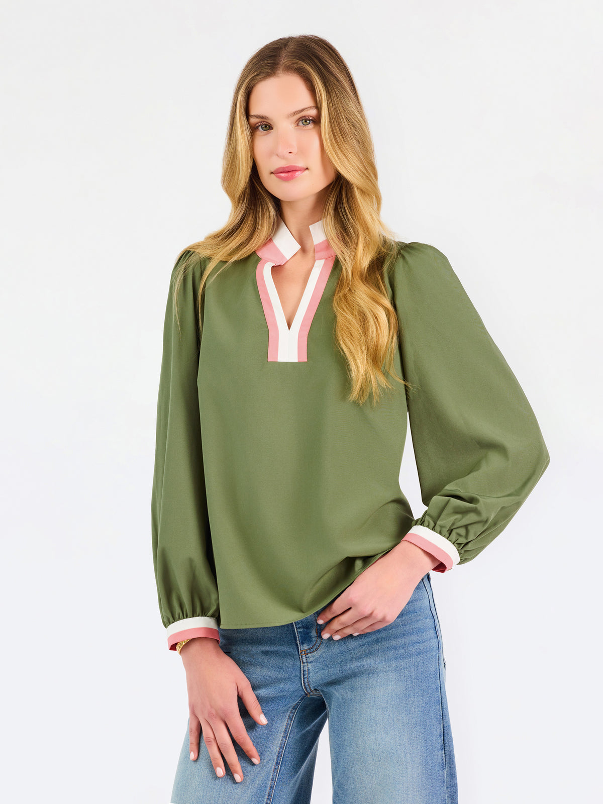 A woman modeling a long-sleeve olive green blouse with a pink and white V-neck trim, paired with blue jeans, against a plain white studio background.