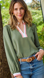 A woman wearing an olive green long-sleeve blouse with a pink and white V-neck placket, paired with blue jeans and a brown belt, standing outdoors next to a tree.
