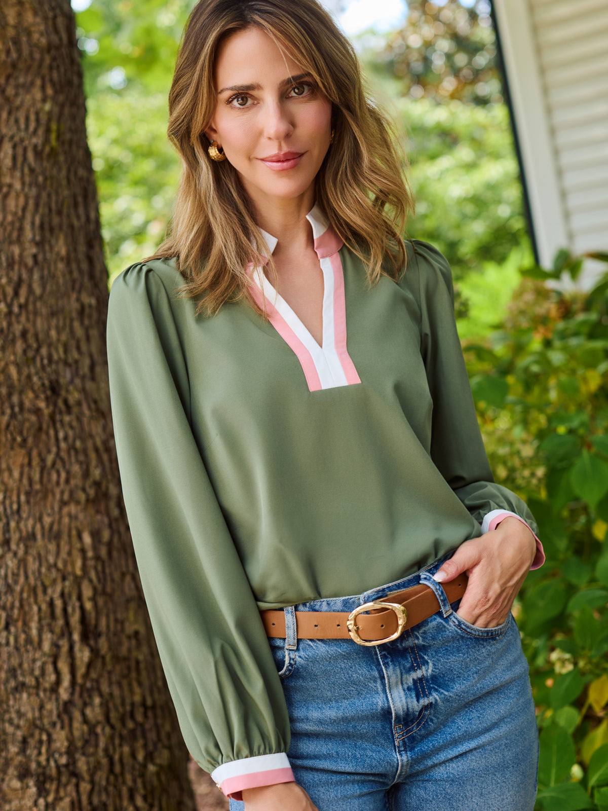 A woman wearing an olive green long-sleeve blouse with a pink and white V-neck placket, paired with blue jeans and a brown belt, standing outdoors next to a tree.