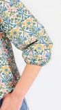 A woman modeling a three-quarter sleeve button-up blouse with a colorful floral and geometric pattern in blue, yellow, and pink, paired with blue jeans against a plain white background.