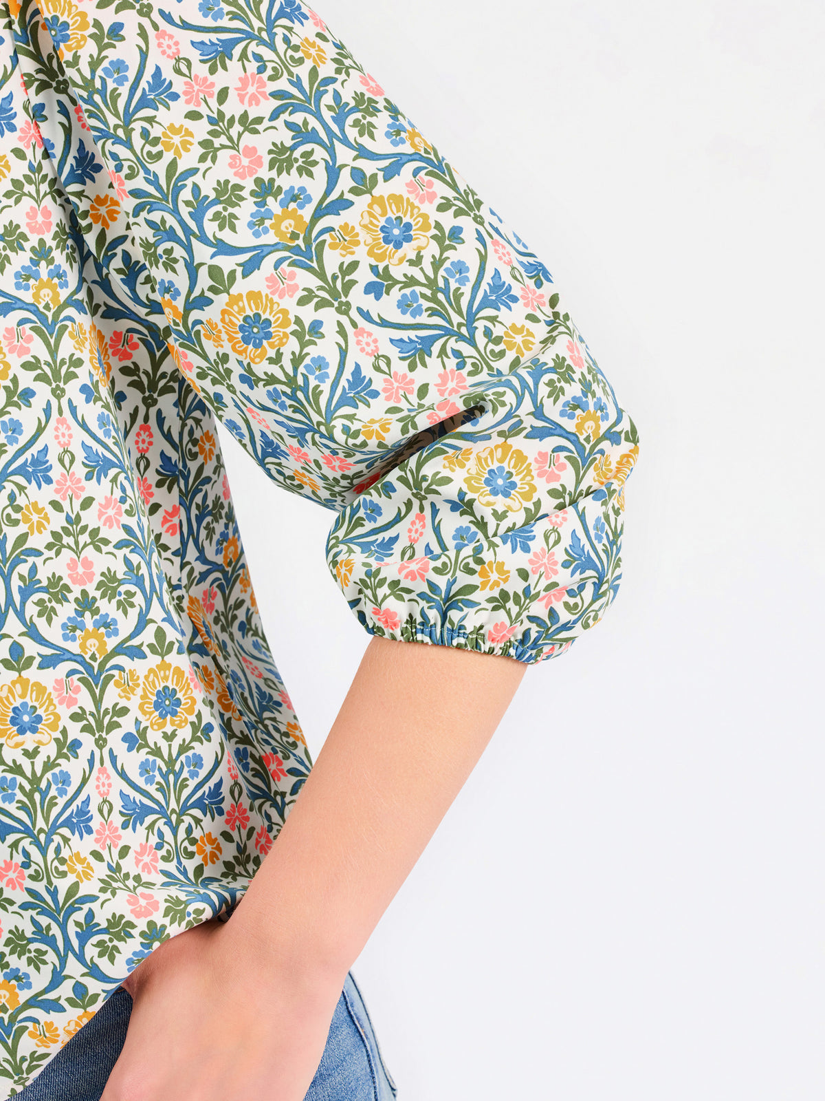 A woman modeling a three-quarter sleeve button-up blouse with a colorful floral and geometric pattern in blue, yellow, and pink, paired with blue jeans against a plain white background.