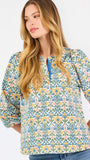 A woman modeling a three-quarter sleeve button-up blouse with a colorful floral and geometric pattern in blue, yellow, and pink, paired with blue jeans against a plain white background.