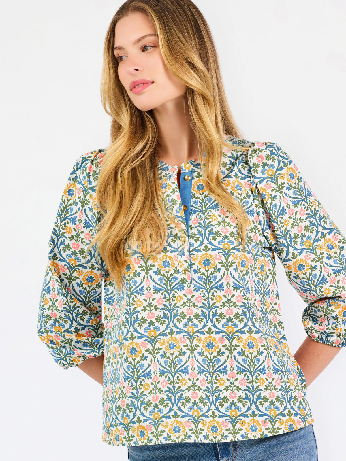 A woman modeling a three-quarter sleeve button-up blouse with a colorful floral and geometric pattern in blue, yellow, and pink, paired with blue jeans against a plain white background.