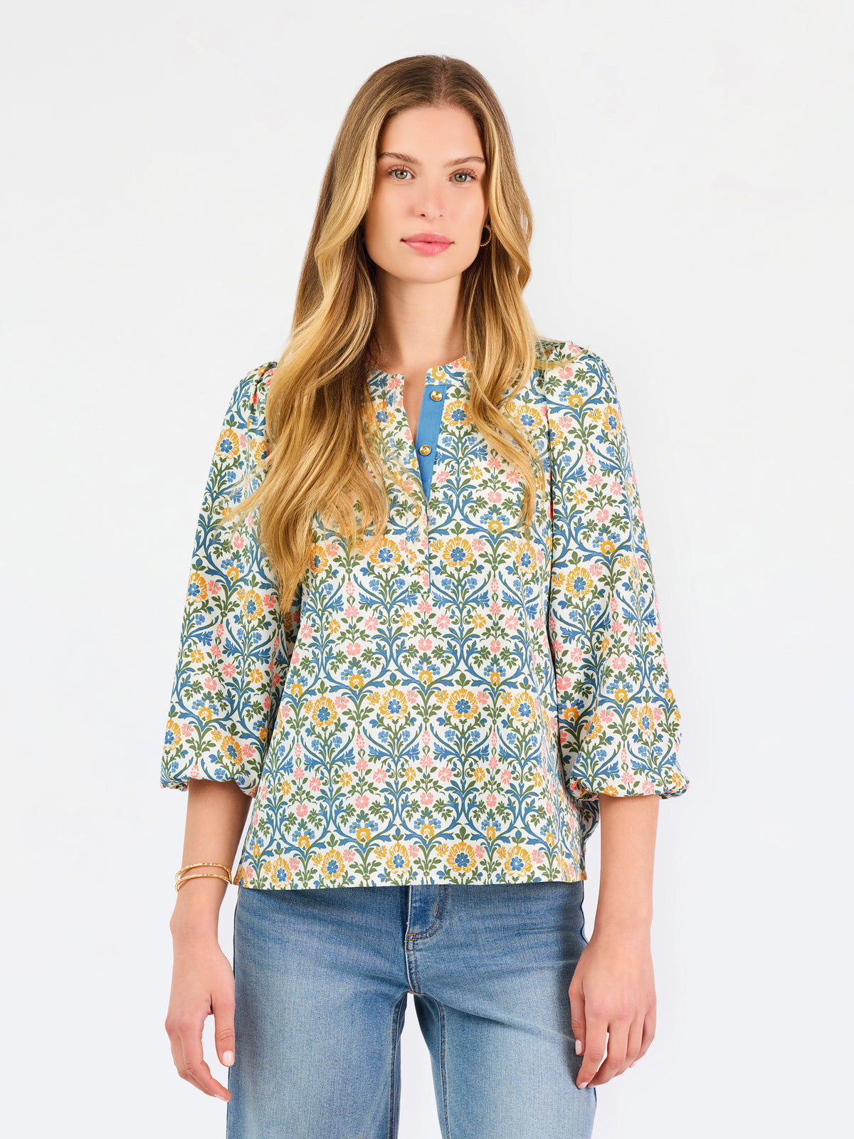 A woman modeling a three-quarter sleeve button-up blouse with a colorful floral and geometric pattern in blue, yellow, and pink, paired with blue jeans against a plain white background.