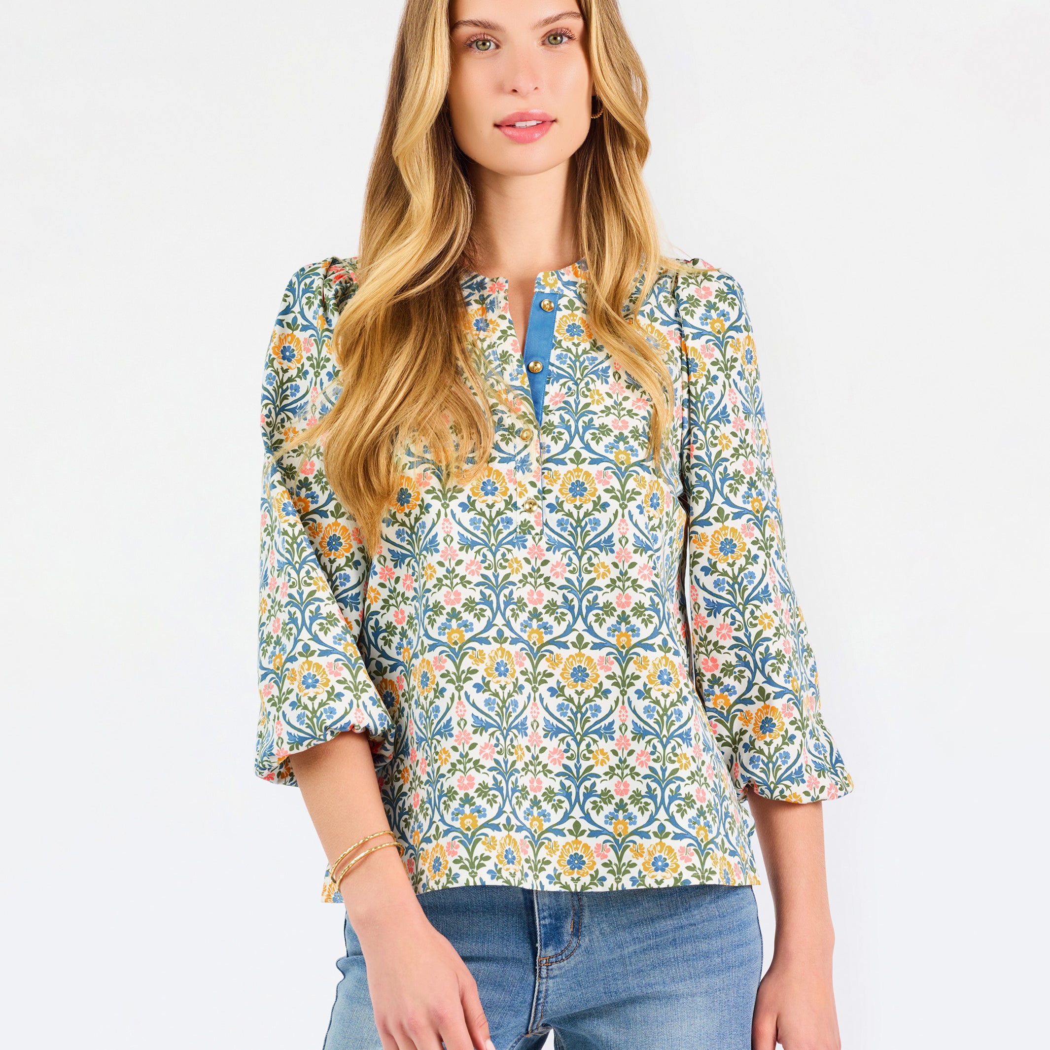  A woman modeling a three-quarter sleeve button-up blouse with a colorful floral and geometric pattern in blue, yellow, and pink, paired with blue jeans against a plain white background.