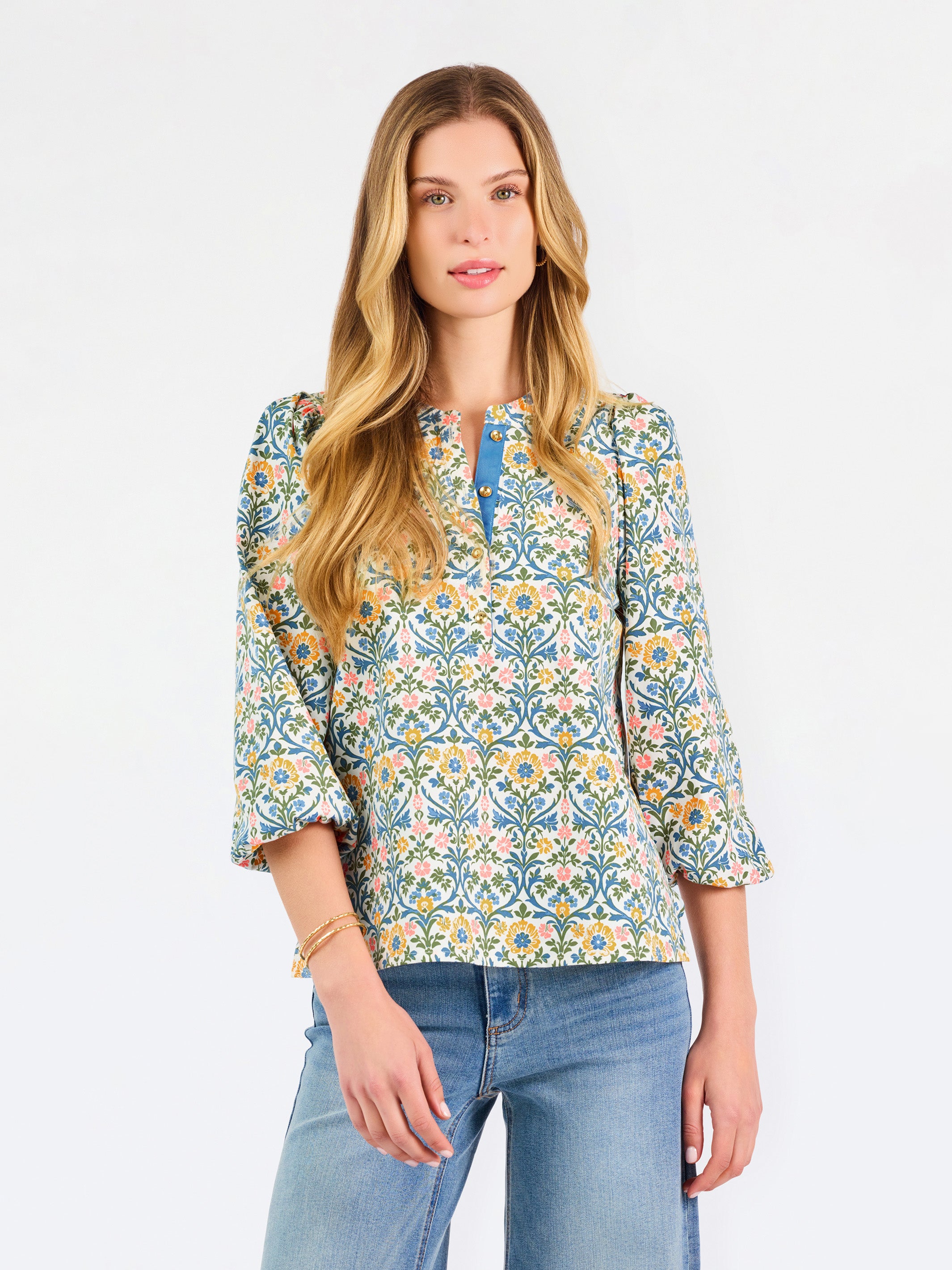  A woman modeling a three-quarter sleeve button-up blouse with a colorful floral and geometric pattern in blue, yellow, and pink, paired with blue jeans against a plain white background.