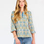  A woman modeling a three-quarter sleeve button-up blouse with a colorful floral and geometric pattern in blue, yellow, and pink, paired with blue jeans against a plain white background.