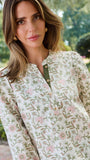 A woman wearing a long-sleeve button-up blouse with a delicate pink and green floral pattern on a cream background, paired with blue jeans, standing outdoors in front of a white column and green trees.